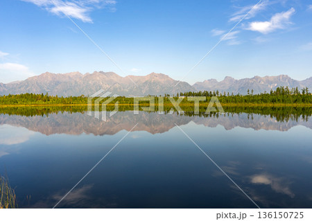 Calm lake with reflections of forest and mountains under a blue sky Calm lake with reflections of forest and mountains under a blue sky 136150725