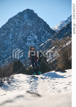Woman photographer hiking a snowy mountain path with winter mountain range background. Adventure travel concept for tourism promotion. Woman photographer hiking a snowy mountain path with winter mountain range background. Adventure travel concept for tourism promotion. 136150727