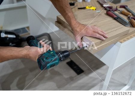 Carpenter drilling a hole in a plywood board using a cordless drill. Furniture assembly process in a home workshop, DIY and woodworking concept. 136152672