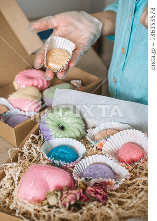 Person holds small soap in paper liner over open cardboard gift box filled with colorful bath products. Handmade bath bombs, personalized self-care gifts, bath salt mixology, aesthetic gift packaging. Person holds small soap in paper liner over open cardboard gift box filled with colorful bath products. Handmade bath bombs, personalized self-care gifts, bath salt mixology, aesthetic gift packaging. 136153578
