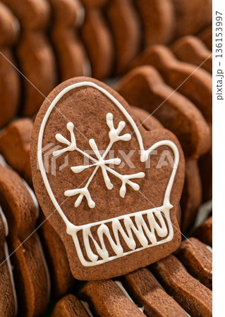 Captured from a macro perspective, a mitten-shaped gingerbread cookie with an intricate white royal icing snowflake design stands out against a background of stacked cookies. The detailed decoration 136153997