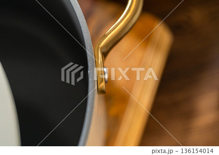 A close up view shows a modern saucepan with a large gold handle resting on a stovetop. The dark interior contrasts with the metallic details. The scene highlights kitchen equipment and cooking tools. 136154014