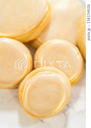 A collection of round sugar cookies decorated with smooth white royal icing and a shimmering gold dust finish. One cookie is being picked up, showing the delicate texture and elegant detailing. These A collection of round sugar cookies decorated with smooth white royal icing and a shimmering gold dust finish. One cookie is being picked up, showing the delicate texture and elegant detailing. These 136154026