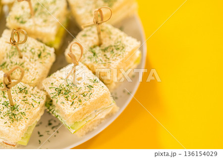 Mini cucumber tea sandwiches arranged on a white plate, placed over a vibrant yellow background. Each sandwich features a layer of sliced cucumber and cream cheese between soft white bread, topped 136154029