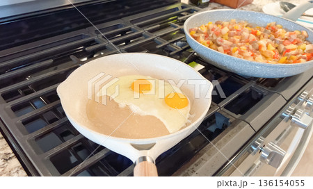 A ceramic pan holds two eggs frying on a stove, with a second skillet of cooked hash in the background. The setup shows a busy breakfast preparation. A ceramic pan holds two eggs frying on a stove, with a second skillet of cooked hash in the background. The setup shows a busy breakfast preparation. 136154055