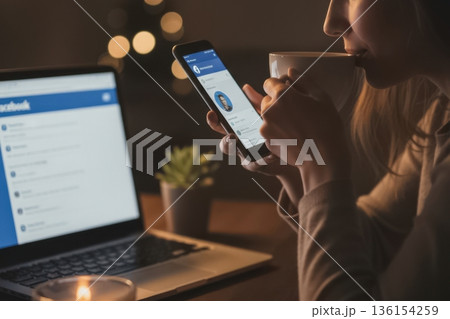 A woman enjoys a cup of coffee while browsing her smartphone beside an open laptop in a dimly lit cozy setting. The background features warm decorative lights, creating an intimate evening mood. The 136154259