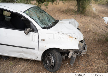 Crashed white car abandoned in dry roadside grass, showing shattered windshield, a missing front bumper, and a flat tire Crashed white car abandoned in dry roadside grass, showing shattered windshield, a missing front bumper, and a flat tire 136154720