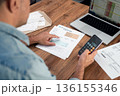 Man calculating household expenses with a smartphone and utility bills on a wooden table. Budget planning and financial management at home. Household budgeting concept. 136155346