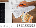 Top view of a person filling out tax forms with an orange pen, euros, and a laptop on a wooden desk. Financial accounting and tax declaration process  136155352
