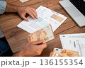 Man calculating utility bills and household expenses at a wooden table. Close-up of hands holding euro banknotes and utility bill papers with a laptop nearby. Household finance management  136155354