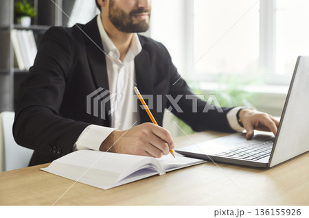 Focused businessman writing notes at desk with laptop in bright office workspace 136155926
