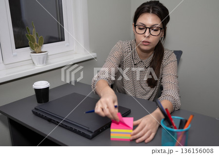 Woman Writing on Sticky Notes While Sitting at a Desk in an Office Space During the Day Woman Writing on Sticky Notes While Sitting at a Desk in an Office Space During the Day 136156008