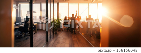 Group of businesspeople celebrating success with arms raised in a modern office with large windows and sunset view, showing teamwork, achievement, and positive work environment 136156048