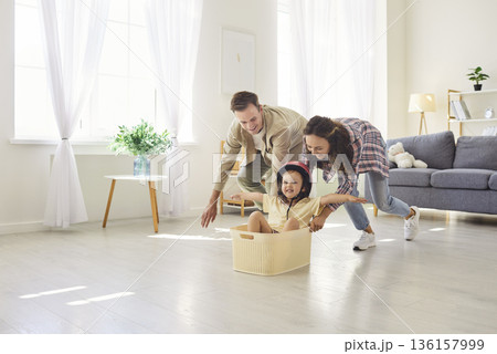 Happy parents push toddler in basket at home, playful family time in living room Happy parents push toddler in basket at home, playful family time in living room 136157999