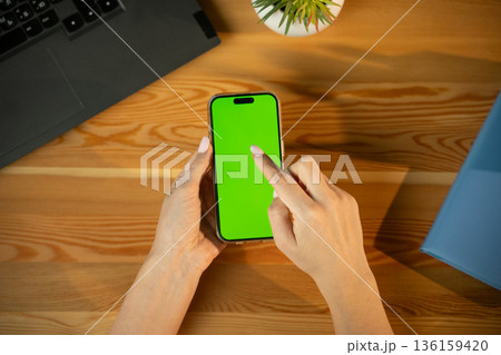 Woman's hands holding a modern smartphone with vivid green chroma key screen, tapping mockup over wooden desk with laptop and plant, top-view workspace for app or ad mockup Woman's hands holding a modern smartphone with vivid green chroma key screen, tapping mockup over wooden desk with laptop and plant, top-view workspace for app or ad mockup 136159420