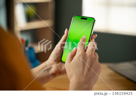 Woman's hands are holding a modern smartphone with a blank green screen. Actively tapping and interacting with the display for a mobile application or digital content. With a blurred office background Woman's hands are holding a modern smartphone with a blank green screen. Actively tapping and interacting with the display for a mobile application or digital content. With a blurred office background 136159425