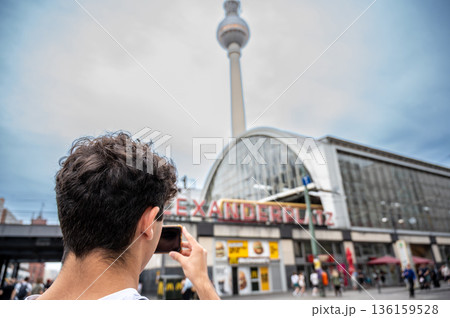 Berlin, germany, august 11, 2023. Young man using a smartphone to capture the iconic fernsehturm and alexanderplatz station, enjoying travel in germany Berlin, germany, august 11, 2023. Young man using a smartphone to capture the iconic fernsehturm and alexanderplatz station, enjoying travel in germany 136159528