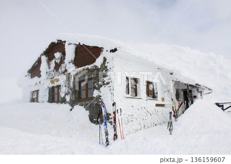 In a winter landscape stands a building covered with snow, surrounded by various skis. 136159607