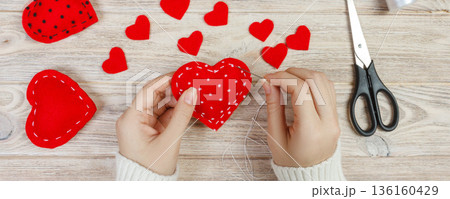 Valentine day theme. Workplace for preparing handmade decorations. Top view of female hands sew felt heart. Packed gifts, tools shabby wooden table 136160429
