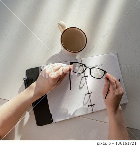Hands holding eyeglasses over an open planner. Light and shadow falling on a notebook, concept of planning and vision 136161721