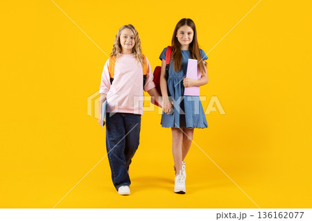 Two preteen schoolgirls walk hand in hand, holding notebooks on yellow background Two preteen schoolgirls walk hand in hand, holding notebooks on yellow background 136162077