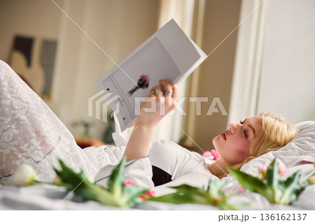 Relaxed woman enjoying fashion magazine, Quiet woman lying in bed with tulips and warm sunlight 136162137