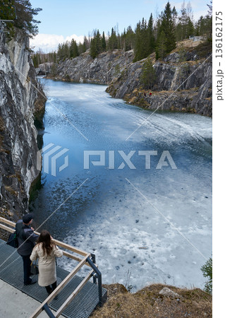 Man and woman enjoying view of partially frozen lake surrounded by rocky cliffs and pine forest in Ruskeala Park, cold weather tourism, travel destination concept Man and woman enjoying view of partially frozen lake surrounded by rocky cliffs and pine forest in Ruskeala Park, cold weather tourism, travel destination concept 136162175