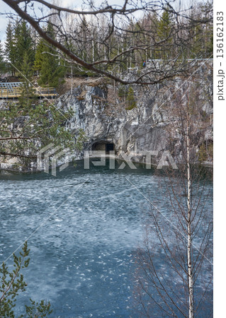 Green tree branches over partially frozen lake with cliff and cave in Ruskeala mountain park in early spring. Nature landscape concept. 136162183