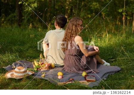 Pair resting back to back beneath trees with picnic and sunset glow Pair resting back to back beneath trees with picnic and sunset glow 136162214