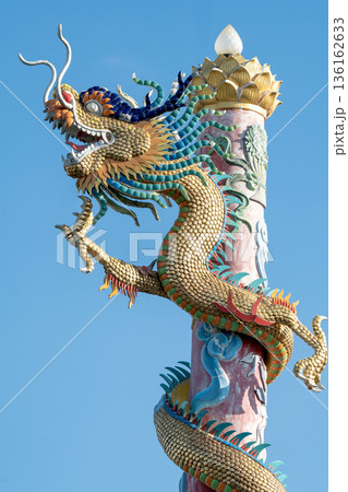 Intricately detailed Chinese dragon statue with golden scales coiled around a tall red pillar. Traditional religious architecture at a temple set against a clear blue sky on a bright sunny day. 136162633