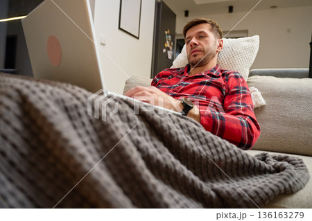 Man in red pajamas lying on sofa under blanket and using laptop Man in red pajamas lying on sofa under blanket and using laptop 136163279