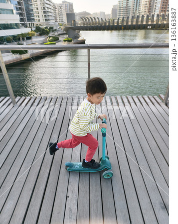 Child Riding A Turquoise Scooter Along A Scenic Waterfront Path. 136163887
