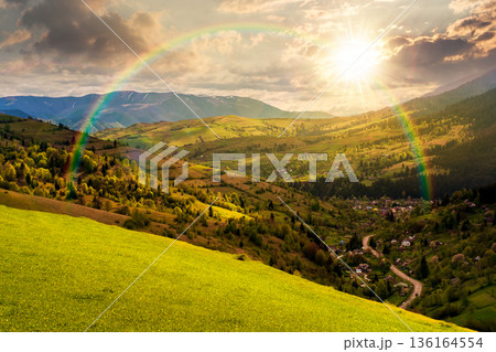 mountainous countryside landscape in spring at sunset. village down in the valley in evening light. grassy rural fields on rolling hills. distant ridge with spots of snow. cloudy weather with rainbow mountainous countryside landscape in spring at sunset. village down in the valley in evening light. grassy rural fields on rolling hills. distant ridge with spots of snow. cloudy weather with rainbow 136164554