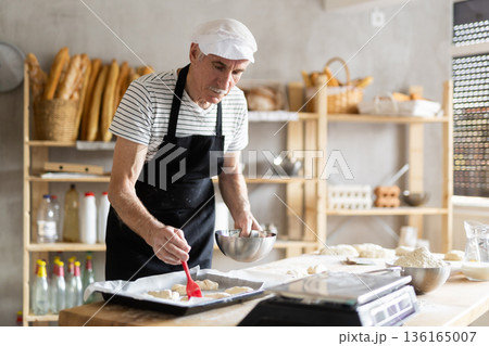 Mature baker brushing uncooked dough pieces on baking sheet with oil before baking in oven 136165007