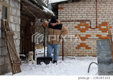 A tired but happy man stands in the yard after clearing snow. A man stands with a snow shovel in a snowy yard and takes a break from winter activities with his small dog nearby, indicating hard work. 136165103