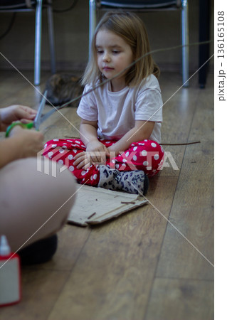 Vertical. A girl sits on a wooden floor and watches intently as her mother snips natural branches with scissors, engaging in creative handicrafts indoors. The family is making a craft. Vertical. A girl sits on a wooden floor and watches intently as her mother snips natural branches with scissors, engaging in creative handicrafts indoors. The family is making a craft. 136165108