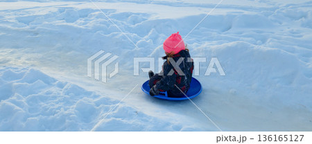 Child enjoying winter sport sitting on a blue sled disc, sliding down an icy path surrounded by snow, engaging in outdoor recreation and childhood fun. 136165127