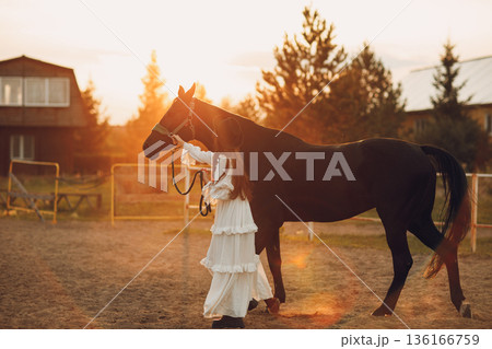 Woman in white dress and black hat with her horse at sunset outdoors ranch 136166759