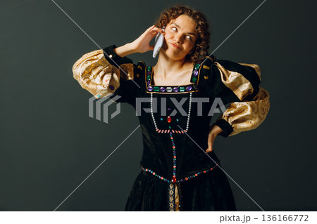 Portrait of a young aristocratic woman talking on mobile phone dressed in a medieval dress on dark background Portrait of a young aristocratic woman talking on mobile phone dressed in a medieval dress on dark background 136166772