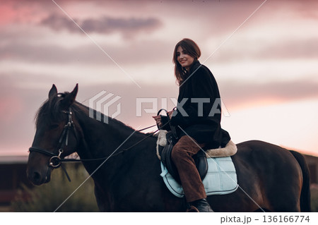 Young woman in black with her horse at sunset outdoors Young woman in black with her horse at sunset outdoors 136166774