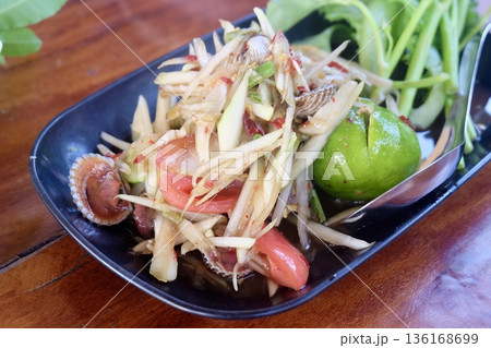 A vibrant close up of spicy Thai green papaya salad with shredded papaya fresh tomatoes chilies and cockles served in a black bowl with vegetables. A vibrant close up of spicy Thai green papaya salad with shredded papaya fresh tomatoes chilies and cockles served in a black bowl with vegetables. 136168699