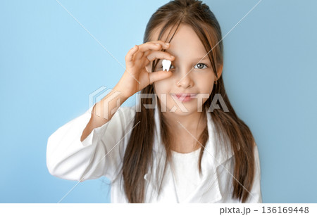 Smiling child holding a lost milk tooth near eye on blue background, symbol of first tooth loss, oral hygiene education, pediatric dentistry and World Oral Health Day awareness on March 20. 136169448