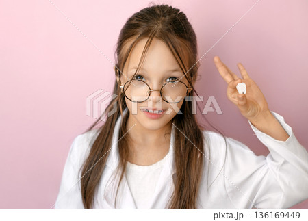 Smiling child in medical coat holding first baby tooth on pink background, concept of National Children Dental Health awareness, milk teeth loss, pediatric dentistry, oral hygiene education and preven 136169449