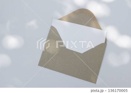 Minimal envelope mockup with blank card on white background in sunlight. Open kraft paper envelope. Invitation, natural stationery flat lay, top view. Minimal wedding, branding template, copy space 136170005