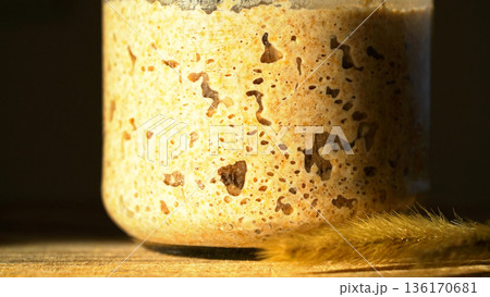 Close-up of fermenting rye sourdough with a bubbly texture, showcasing wild yeast in home baking. Starter in a glass jar with wheat stalk, demonstrating active yeast. Healthy fermented food commercial Close-up of fermenting rye sourdough with a bubbly texture, showcasing wild yeast in home baking. Starter in a glass jar with wheat stalk, demonstrating active yeast. Healthy fermented food commercial 136170681