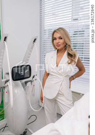 Professional female cosmetologist working in contemporary medical aesthetic clinic, standing confidently by advanced SMAS lifting equipment for facial rejuvenation and body contouring procedures. 136172640
