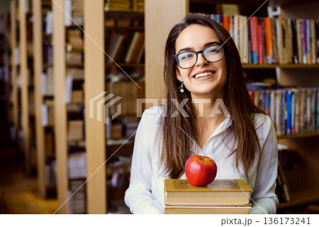 Happy female student with pile of books in the library. Smiling attractive girl eager to study and get new knowledge 136173241