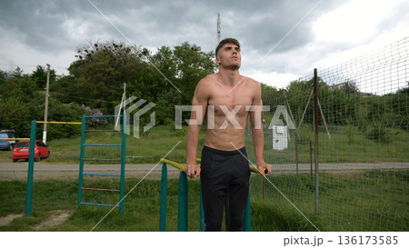 Young sportsman doing push ups on parallel bars at nature. Strong sporty man exercising at sports ground. Male shirtless athlete training hard outdoor. Concept of active sportive lifestyle. Slow mo Young sportsman doing push ups on parallel bars at nature. Strong sporty man exercising at sports ground. Male shirtless athlete training hard outdoor. Concept of active sportive lifestyle. Slow mo 136173585