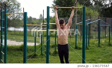 Muscular athlete doing pull ups on horizontal bar at sports ground. Young shirtless sportsman exercising at nature. Strong sporty man training outdoor. Concept of healthy and active lifestyle. Slow mo 136173587