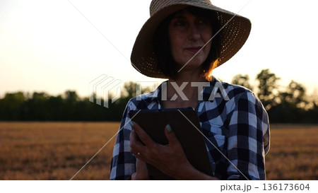 Female agronomist in straw hat monitoring harvest on barley field at sunset. Adult farmer using digital tablet on wheat meadow at dusk. Beautiful scenic landscape. Concept of agricultural business Female agronomist in straw hat monitoring harvest on barley field at sunset. Adult farmer using digital tablet on wheat meadow at dusk. Beautiful scenic landscape. Concept of agricultural business 136173604